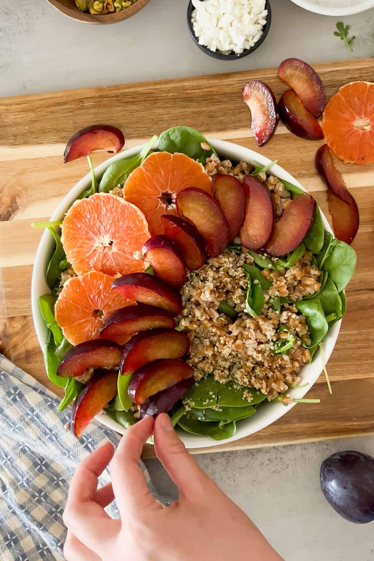 top down shot of assembling salad; adding sliced plums and oranges, farro and hemp seeds to mixed spinach and arugula in bowl